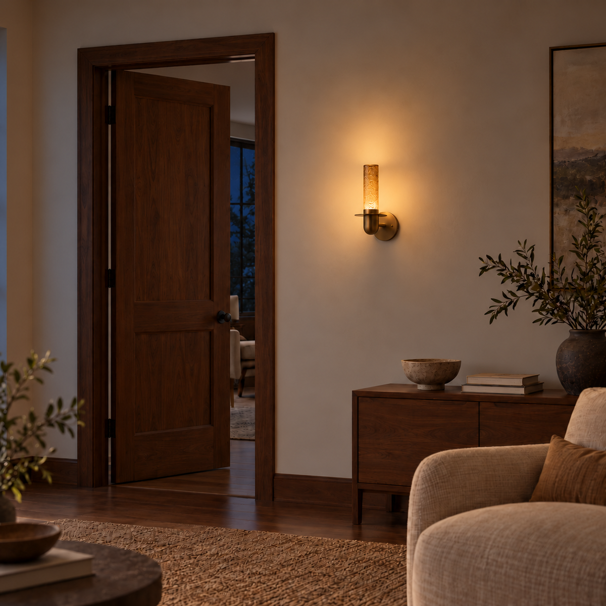 Textured tea glass cylinder wall light illuminated above a wooden cabinet beside an open doorway and upholstered chair at night.